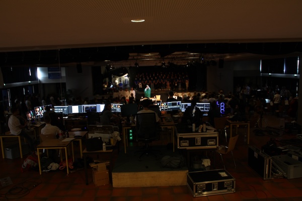 Mischpulte, Monitore und Menschen von hinten, im Hintergrund eine Bühne mit Chor, bunt beleuchtet